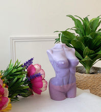 Load image into Gallery viewer, A purple candle in the shape of a female torso, placed on a table with decorative flowers and a green plant in the background.
