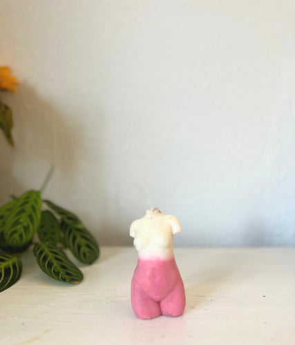 Small female torso candle with a pink base and white top on a light surface with a blurred plant in the background.