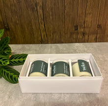 Load image into Gallery viewer, Three candles in a white box on a concrete surface with a wooden wall in the background.
